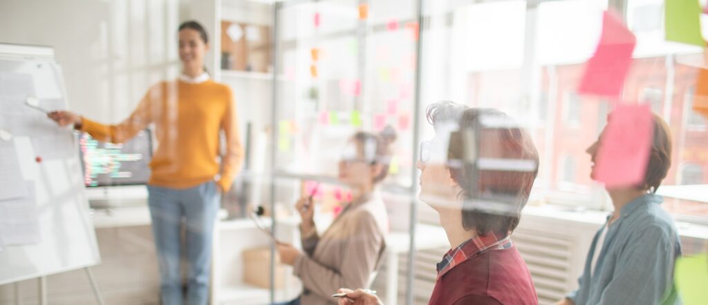 Seminarteilnehmende sitzen hinter einer Glasscheibe und hören der stehenden Referentin zu.